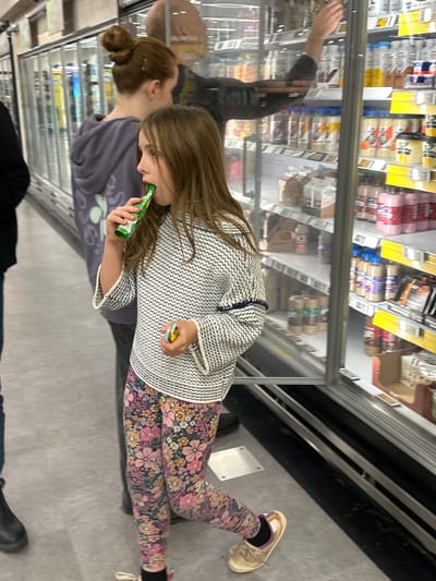 The image depicts a young Caucasian girl with long brown hair, wearing a patterned sweater and floral leggings, consuming a green juice box in a grocery store. She is standing in front of a refrigerated aisle filled with various beverages. To her left, a Caucasian woman with her hair in a bun, wearing a grey hoodie, stands behind the girl. The background reveals shelves stocked with numerous bottles and containers, indicative of a retail food environment. The flooring appears to be concrete or linoleum. The lighting suggests an indoor, commercial setting. The image was taken in the city of Toormina, Australia, a suburb of Coffs Harbour.