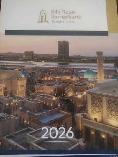The image shows a brochure for the "Silk Road Samarkand Tourist Centre" in Bahshitpa, Uzbekistan. The brochure features a nighttime aerial view of a modern tourist complex with traditional architectural elements. In the foreground, there are several multi-story buildings with flat roofs, adorned with intricate geometric patterns. Some buildings have illuminated windows, and several domes are visible, including a prominent turquoise dome. A tall, slender tower with decorative patterns stands to the right. In the background, a river or large body of water is visible, with a modern high-rise building (labeled "Hilton") and other structures on the opposite bank. The sky is a soft twilight orange. Large white numerals "2026" are superimposed on the lower right portion of the image. There are no people clearly visible in the scene.