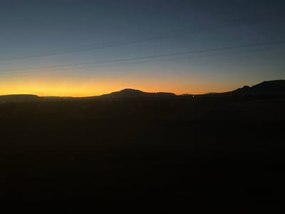 A dusky landscape features distant mountains silhouetted against a gradient sky transitioning from deep blue to a vibrant orange at the horizon. A flat, dark foreground stretches towards the mountains, obscuring details of the terrain. Power lines are visible in the upper part of the image. The scene evokes a sense of peaceful vastness, captured during the early morning or late evening hours.