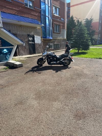 A black motorcycle is parked on a gravel lot in front of a brick building. A person wearing a dark shirt and shorts stands next to the motorcycle. The building has a blue-tinted glass facade and a blue metal roof over an entrance. A green spruce tree is visible behind the building. The scene is illuminated by bright sunlight, casting shadows on the ground. The location is likely in a residential area of Mariinsky, Russia.