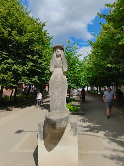 The photograph depicts a sunlit outdoor scene in Bucha, Ukraine, on a warm day with scattered clouds. The foreground is dominated by a weathered stone sculpture of a woman with a floral wreath around her head, her hands covering her ears. The sculpture is positioned on a concrete pedestal.
To the left of the sculpture, a paved walkway lined with mature trees leads into the background. An elderly woman, dressed in a white shirt and patterned pants, walks away from the camera, carrying a bag. Further back, other people are visible in the distance, walking along the path. Buildings, possibly shops or residences, are partially visible behind the trees.
To the right of the sculpture, a man in a gray t-shirt and shorts walks towards the camera. His hands are in his pockets, and he appears to be mid-stride. Sunlight casts dappled shadows from the trees onto the walkway. The environment suggests a public space, likely a park or a pedestrian thoroughfare within the city.