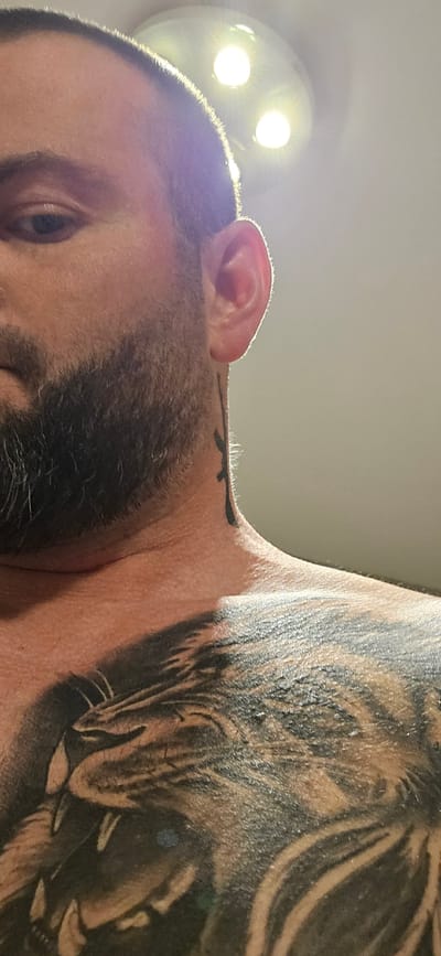 A close-up, low-angle photograph depicts the face and upper torso of a light-skinned man. The man has dark hair, stubble, and a beard. His right eye is visible. A tattoo of a wolf's head adorns his left shoulder and upper chest. Part of his ear is visible to the right. A light fixture with two illuminated bulbs is in the upper right corner of the image. The location in the city of Gütersloh, Germany, cannot be determined from the image.