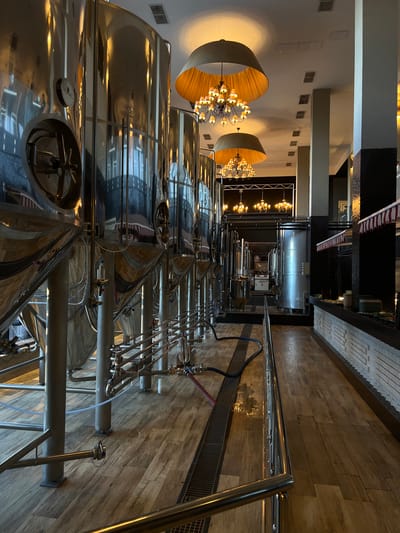 The image captures a wide-angle view of a brewery's fermentation room, likely in Lesnoi Gorodok, Russia. The scene is dominated by numerous large, polished stainless steel fermentation tanks arranged in rows. These tanks are connected by an intricate network of pipes, valves, and gauges, indicating an active brewing process. Above, a high ceiling with industrial lighting and decorative chandeliers creates a contrast between the functional brewing equipment and the establishment's aesthetic.
The floor is made of wooden planks, and a stainless steel railing runs along the right side of the image, suggesting a viewing or access area. The lighting in the room is warm and ambient, reflecting off the shiny surfaces of the tanks, which creates a somewhat dramatic and upscale atmosphere. There are no people visible in the image. The time of day is not explicitly clear, but the artificial lighting suggests it could be evening or a time when artificial light is preferred. No weather cues are present as the image is indoors.
Notable details include the large, circular wooden light fixtures hanging from the ceiling, providing a warm glow. The reflections on the stainless steel tanks show glimpses of the surrounding architecture and lighting. One of the tanks has a small circular plate with embossed text that appears to read "MOMAC". The overall impression is a modern, well-maintained brewery with a focus on both functionality and design. The activity taking place is clearly the process of fermentation, a crucial stage in beer production.