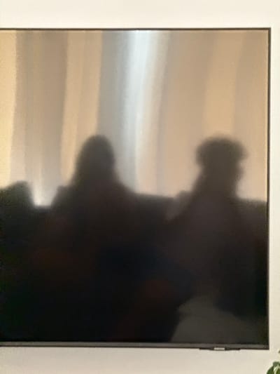 A dark, shadowy image captured on a television screen shows the silhouettes of two individuals sitting on a couch. The background appears to be a softly lit curtain, with vertical lines of light and shadow. The individual on the left is larger, with a rounded head, while the individual on the right is slightly smaller and appears to be wearing a cap. The scene is dark, with the details of the people and their surroundings obscured by shadow, giving the impression of a dimly lit room or evening viewing. This image was taken inside a residence in Mesogi, Cyprus.