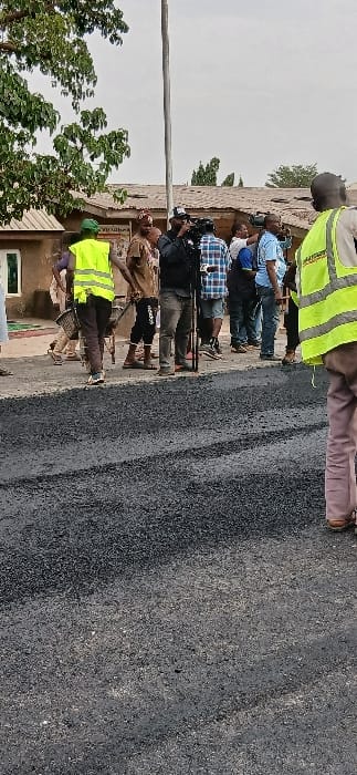 A group of people gathers along a newly paved road. Several wear casual clothing, while two individuals sport bright yellow safety vests. A video camera on a tripod is visible amidst the group. Trees and buildings border the scene, and a pole stands vertically to the left. The asphalt road surface is dark, contrasting with the lighter-colored sky.