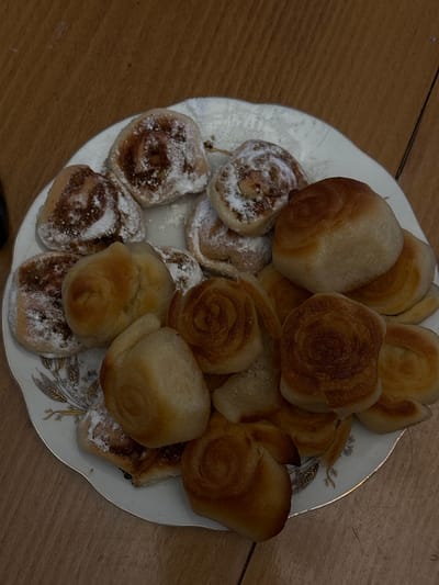 A plate of pastries sits on a wooden surface. Some pastries are round and sprinkled with powdered sugar, revealing a brown swirl pattern. Other pastries are smooth, golden-brown rolls. The plate itself is white with a decorative gold-colored pattern around the rim. The image is taken from an overhead perspective. There is no indication of a location in Borino, Bulgaria.