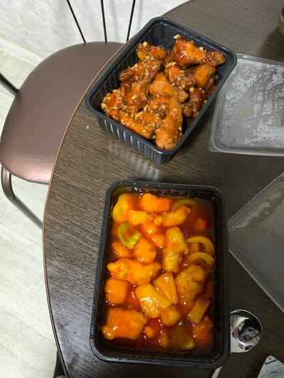 Two black rectangular takeout containers of food sit on a dark wood-grain surface, possibly a table. The container on the left holds several pieces of fried chicken coated in a glossy, amber-colored sauce and sprinkled with chopped garlic. The container on the right contains pieces of fried food, likely chicken or fish, coated in a red sweet and sour sauce along with chunks of pineapple and slices of bell pepper, which appear to be yellow and green. A metal spoon with a reflective surface is visible to the right of the right container, and its reflection shows part of a ceiling and possibly a person. The overall lighting suggests an indoor setting, possibly at home, with no clear indication of the time of day or weather. There is no visible text in the image. The scene depicts a meal being served or ready to be eaten.
