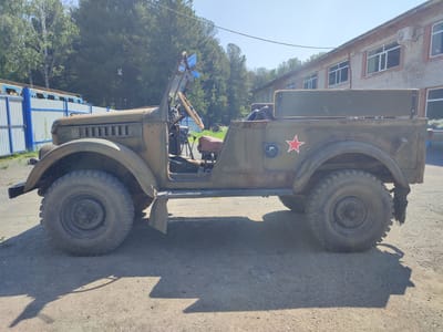 A weathered, olive-drab, Soviet-era military jeep, possibly a GAZ-67 or similar model, is parked on a dirt and gravel surface in Zorkaltsevo, Russia. The jeep is viewed from its left side, showcasing its open cockpit with a brown vinyl seat visible. A red star insignia is painted on the rear passenger side panel. The vehicle has large, rugged tires and a spare tire mounted on the rear.

Behind the jeep, a light-colored brick building with multiple windows and an air conditioning unit is situated. To the left of the jeep, a light blue and white fence runs parallel to a blue and yellow structure. Tall green trees form a backdrop against a clear blue sky. The overall scene suggests an outdoor setting, possibly near a storage facility or an industrial area. The lighting indicates it is daytime, with the sun casting distinct shadows from the jeep onto the ground. There are no people visible in the image.