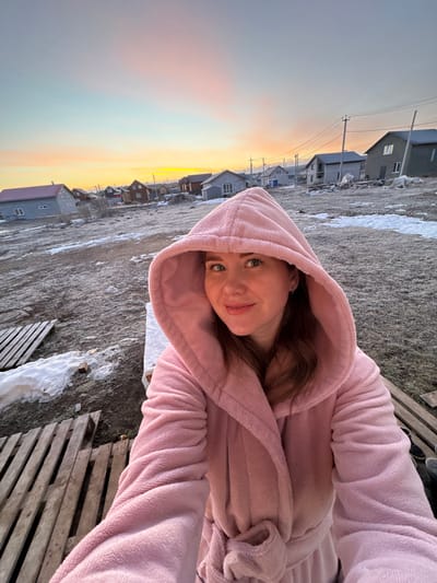 A woman in a pink hooded robe is facing the camera. The setting is Nestiukovo, Russia, recognizable by the style of buildings in the background and known geographic characteristics. Snow patches are present on the ground, suggesting a cold climate. A wooden pallet is located on the right. The sky shows a sunset with a gradient of colors, ranging from yellow to grey.