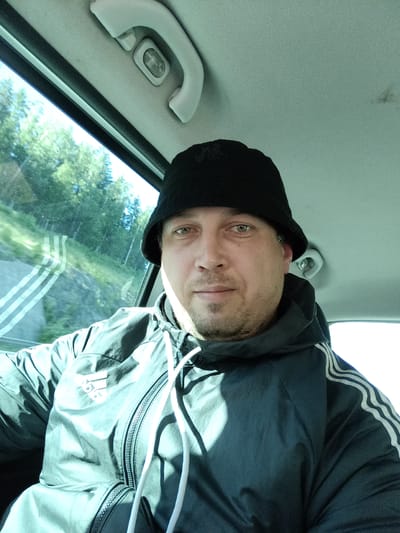 A man is taking a selfie inside a vehicle. He is wearing a black bucket hat and a dark jacket with white stripes on the sleeves. The man has light skin and light-colored eyes, and is looking directly at the camera. Through the window, a wooded area with a road is visible. The location is Drahňovice, Czechia.