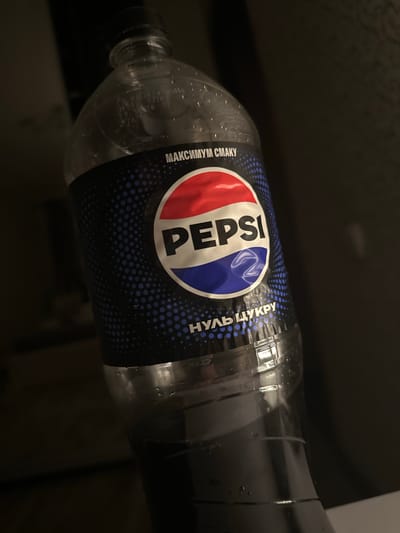 The image shows a bottle of Pepsi with a black label. The label has the Pepsi logo and text that appears to be in Cyrillic. The text "МАКСИМУМ" and "НУЛЬ ЦУКРУ" are visible. The bottle is partially filled with a dark liquid and has condensation on the outside. The background is dark and out of focus, suggesting an indoor setting. There is no discernible location information within the image that would place it in Podilsk, Ukraine, or any other specific geographical area.