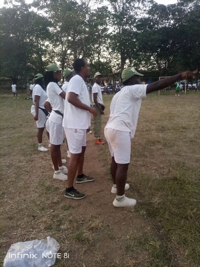 This outdoor image captures a group of people, likely National Youth Service Corps (NYSC) members, participating in an event on a grassy field in Ayere, Nigeria. The time of day appears to be late afternoon or early evening, indicated by the soft, warm light and the slightly overcast sky visible above the trees.

The primary subjects are several individuals, mostly young adults, dressed in white T-shirts, white shorts, and green caps, forming what looks like a line or formation. Many also wear white or green-striped socks and various sneakers, some with visible Nike branding. One man in the foreground on the right side of the frame is prominently featured, extending his right arm to point towards something out of frame to the right, suggesting he is giving instructions or drawing attention to an activity. Another person further back on the left also appears to be pointing in a similar direction.

The ground beneath them is a mix of dry grass and dirt. In the background, numerous trees line the perimeter of the field. Beyond the immediate subjects, other people are visible further down the field, some standing, others possibly seated or observing, suggesting a larger gathering or ongoing event, possibly a sporting match like football given the general layout of the field and distant figures. One individual in the mid-ground on the right wears camouflage-patterned trousers, distinct from the white uniform, perhaps an instructor or official.

A clear plastic bag is visible on the ground in the bottom-left corner of the image. The text "Infinix NOTE 8i" is watermarked in the bottom-left corner, indicating the device used to capture the photo.