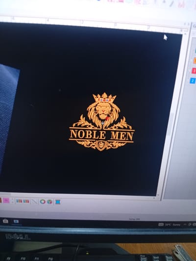 The image shows a computer monitor displaying an embroidery design of a lion wearing a crown, with the text "NOBLE MEN" below it. The design is rendered in gold and red tones against a black background. The monitor is part of a computer setup, with a keyboard visible at the bottom of the frame. The computer's operating system taskbar is visible at the very bottom, showing icons and system information. The environment appears to be an indoor setting, likely a home or office, with no specific visual cues to pinpoint a location within Rumuepirikom, Nigeria.