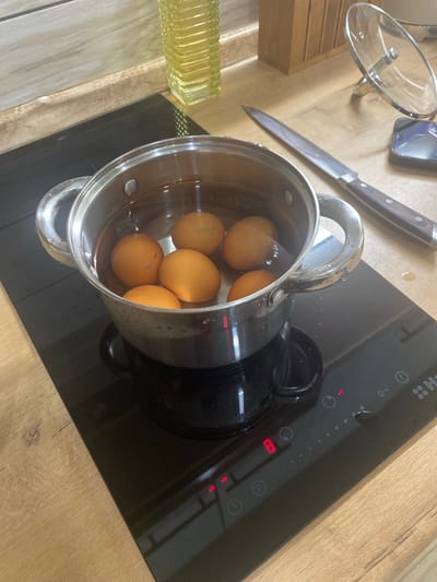 This image captures a domestic kitchen scene in the city of Копнино, Russia. A stainless steel saucepan containing six brown eggs submerged in water is positioned on the front-left burner of a black induction stovetop. The stovetop's integrated control panel, located to the right of the pot, displays the number '8' in red LED, indicating the heat level, alongside various touch-sensitive icons for power, timer, and a child lock, and a partial "HA..." branding. Small water droplets are visible on the stovetop surface and the exterior of the saucepan, suggesting active boiling or condensation.
The countertop, made of a light brown wood-grain laminate, extends across the scene. To the right of the stovetop, a long chef's knife with a dark wooden handle and a silver blade rests horizontally on the countertop, pointing left. Further to the right, a clear glass pot lid with a silver knob is propped upright, leaning against an unseen object or the wall. A dark-colored smartphone lies flat on the countertop adjacent to the lid. In the background, a light-toned, wood-grained backsplash is visible, above which sits a tall, yellow, ribbed glass bottle, possibly an oil dispenser. A wooden block, potentially a knife holder, is also partially visible behind the lid and smartphone.