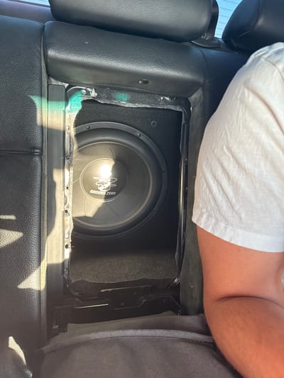 The image depicts an in-car audio system installation. A Ground Zero subwoofer is mounted in an enclosure situated between the driver and passenger seats, as indicated by the armrest. The upholstery and interior suggest the scene is within a vehicle. No direct evidence places the scene in Manolsko Konare, Bulgaria. No actions or interactions are visible. A person's arm and shoulder, wearing a white shirt, are partially visible in the top of the image.