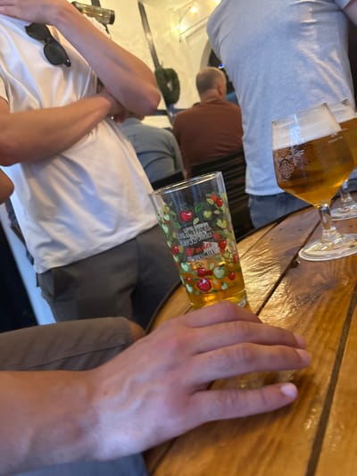 A hand rests on a wooden table with two glasses of beer on it. One glass is filled with an amber liquid, the other is a tall glass with a colorful design of red and green apples and the words "LOCAL APPLES BROWN'S FARM OUR MILL" printed on the side, also containing an amber liquid. The table is in a pub, there are two people out of focus in the background, with the left-side person wearing a white shirt, and the right-side person wearing a gray shirt. The scene is located in Ware, United Kingdom at latitude: 51.811, longitude: -0.033.