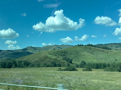 The image shows a landscape with rolling green hills under a bright blue sky dotted with fluffy white clouds. A vast green meadow, possibly a field of wildflowers, occupies the foreground. A line of trees runs along the base of the hills, with more scattered trees and bushes dotting the slopes. The scene appears to be a natural landscape, likely in a rural or mountainous area. The description of Verkh-Aya, Russia, suggests a specific geographical location for this scenery. There are no people or animals visible in the image, and no actions or interactions are depicted. The environment is characterized by natural elements: sky, clouds, hills, grass, and trees.