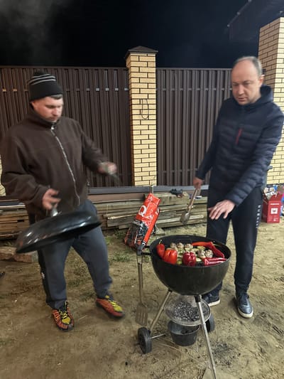 A nighttime scene in слобода Старая, Russia, shows two men standing near a charcoal grill. One man, wearing a dark jacket and dark pants, stands over the grill holding tongs. The grill is filled with food, including red peppers and what appear to be mushrooms or meatballs. The other man, wearing a brown jacket, dark pants, and a beanie, is bending over and holding the lid of the grill in his hand. Bags of charcoal and cooking utensils are visible near the grill. The setting appears to be a backyard or outdoor space near a brown metal fence and a brick structure.