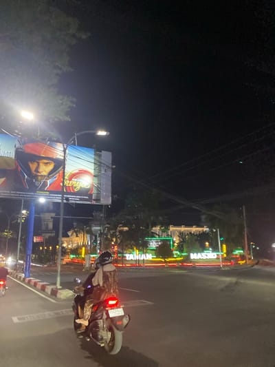 This media file captures a nighttime scene in Macege, Indonesia, likely on a street. A person is riding a motorcycle, seen from the side, wearing a helmet and a patterned jacket. The motorcycle has a license plate that reads "DP-2348 LU". The road is wet, suggesting recent rain, and illuminated by streetlights and bright neon signs.

Prominently displayed are colorful neon signs that spell out "MASJID" and "TAVIAN" vertically. Adjacent to these, a large billboard features an advertisement with the text "KITA ADA SURYA" and a warning about the dangers of smoking, including the phrase "PERINGATAN: MEROKOK SAYA TERKENA KANKER TENGGOROKAN LAYANAN BEER NAMERONOK (2017) DLARANG MENJUAL DAN MEMBERI KEPADA ORANG DI BAWAH 21 TRAIN N MENMAHAL".

The overall impression is a vibrant and somewhat bustling street scene at night, with a mix of traffic, advertising, and architectural lighting. The time of day is clearly night, indicated by the dark sky and artificial illumination. The wet road adds a specific atmospheric detail.
