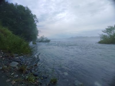 This media file depicts a wide, fast-flowing river under a cloudy, overcast sky. The scene is set along the riverbank, which is lined with dark, wet rocks and lush green vegetation, including tall grass and trees. The water itself is a deep, murky blue-gray, with visible ripples and turbulence, suggesting a strong current.
In the distance, across the river, there is a misty haze obscuring the far bank and any distant mountains or hills. The mist softens the landscape, creating a sense of depth and atmosphere. The overall mood of the image is serene and perhaps a little somber, possibly due to the muted colors and the cloudy weather.
The time of day appears to be early morning or late afternoon, as indicated by the soft, diffused light and the gray sky. The weather is likely cool and damp, given the mist and the dark, wet appearance of the riverbank.
The location is Psebai, Russia, a region known for its natural beauty and rivers. The image captures a moment of tranquility in a natural setting, with the powerful flow of the river being the dominant element. There is no visible text in the image.