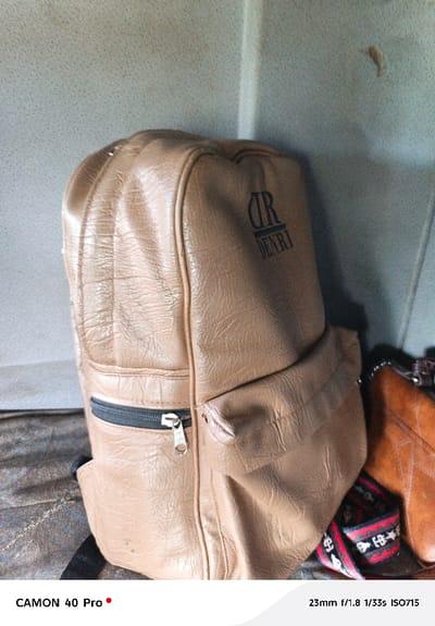 The scene depicts a static arrangement of personal items within what appears to be an interior space in Hells Gate ward, Kenya. A prominent tan, faux-leather backpack, exhibiting creased texture, stands upright in the center-left. It features a visible front pocket with a black zipper and a silver-colored pull tab. A dark, stylized "DR DENRI" logo is present on the upper front of the backpack. To the right of the backpack, a section of another brown bag, possibly a purse, is visible, partially obscured. A strap with a red, black, and white geometric pattern lies between the two bags. All items rest on a dark, textured surface, likely fabric or worn leather. The background consists of a plain, light grey or off-white wall or panel, showing slight discoloration and a small, dark, irregular mark in the upper right portion. No individuals are present.