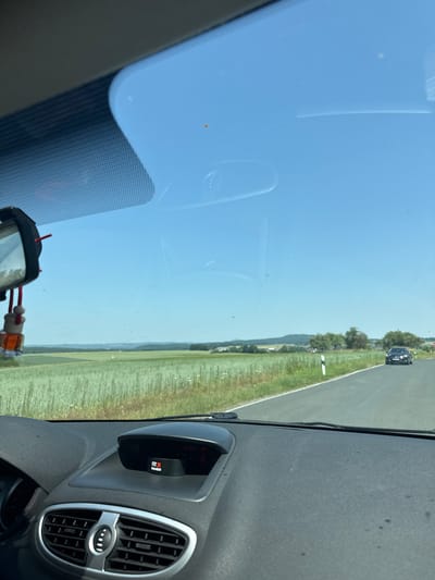 The image shows a view from inside a car driving on a road in Niederwetz, Germany. The dashboard is visible in the foreground, with a car accessory displaying "FIT & MEMBER" on top of it. Through the windshield, a green field stretches to the horizon, with trees and hills in the distance. The sky is clear blue. A dark-colored car is driving on the road ahead.
