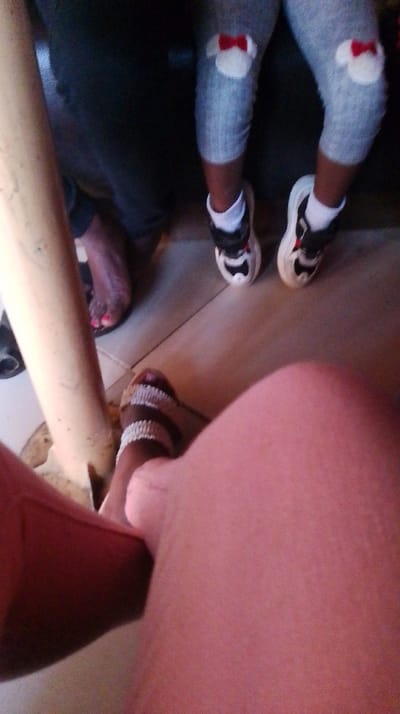 The image shows a low-angle shot of four pairs of legs and feet. The person closest to the camera is wearing a white and brown sandal. To the left of this leg is another person wearing a black sandal with pink nail polish. At the top are child's legs wearing grey leggings with Mickey Mouse patches on the knees and white, black, and red sneakers. A white support pole is also visible. The focus of the photo is on the person with white and brown sandals.