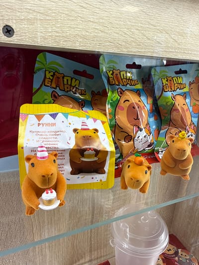 The image, taken in Krilovo, Russia, shows a display of plush toys and packaged confectionery items, likely from a store or vending machine. The scene is viewed from above, looking down into a display case featuring a light wood-grain shelf or surface and a clear glass shelf above.

The primary subjects are three small, flocked, orange-brown plush toys resembling capybaras, arranged across several packages. The top toy has a small yellow duck on its head. The middle toy has a small orange carrot-like accessory. The bottom toy, positioned on a yellow package, wears a striped pink and white party hat and holds a miniature white cupcake.

Behind the toys are several packaged items. Three blue-themed packages feature a cartoon illustration of a capybara lounging in foliage near water, with a white rabbit character beside it. Text on these packages includes "КЭТРИН" or "КЭПРИК" and "Рчик" (likely part of a brand or character name, possibly "Kaprik"). The bottommost package is yellow and features a cartoon bear wearing a party hat and holding a cake. Visible Russian text on this package reads "РУННИ" (Runni), "Кулинар-кондитер. Очень любит сладости. Именинник, човник качества." (Culinary confectioner. Loves sweets very much. Birthday boy, a master of quality.). Additional partial text on this package is visible but fragmented.

To the right, a clear plastic cup with a lid is lying on its side. The lid has "CAUTION: HOT" embossed on it. Behind the cup, several circular, red-themed tokens or coasters with cartoon faces are visible. A black screw is visible on the wooden surface in the bottom-left corner of the image. The lighting is bright and even, typical of an indoor display.