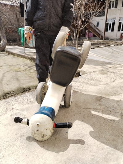 An outdoor scene in Limbur, Uzbekistan, depicts a partial view of an individual standing on an uneven, light-colored concrete surface under direct sunlight. The person, whose lower torso and legs are visible, wears a dark quilted jacket with a white logo on the left sleeve, dark sweatpants, and light-colored gloves, one of which has an orange wristband. They are holding a white, blue, and black four-wheeled child's ride-on toy, oriented with its black seat and rear wheels near the person's legs and its front section, featuring two black handlebars, extending away.

In the background, to the left, are bare deciduous trees, a clothesline with green and white fabric hanging, and a low concrete curb. To the right, a two-story light-colored building with multiple windows and an external wooden staircase is visible, alongside more bare trees. The ground surface shows cracks and patches of dirt, indicating a residential or communal outdoor area. Shadows are distinct on the ground, suggesting bright daylight.