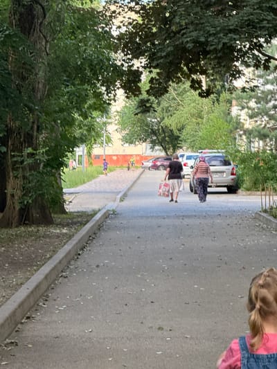 In Ekoposelok, Russia, two women are walking down a paved road lined with trees. One woman, wearing a striped shirt and a patterned skirt, walks ahead. Behind her, another woman in a dark shirt and a light-colored skirt carries a patterned bag. They are walking away from a silver car parked on the side of the road. The scene appears to be outdoors on a sunny day, possibly in the late afternoon or early evening, given the shadows. The trees suggest a residential or park-like setting. The overall mood seems casual and ordinary, with no strong emotional cues. There is no visible text in the image.