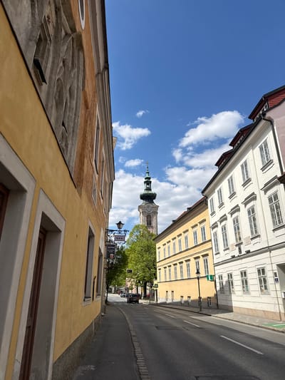The image presents a sunlit street scene in Hainburg an der Donau, Austria, viewed looking along a street lined with historic buildings. A clear blue sky with scattered white cumulus clouds dominates the upper portion, indicating bright, clear weather during the day, likely mid-day.
On the left side of the street, a multi-story building with a warm ochre-yellow facade is prominent in the foreground. This building features intricate, sculpted stucco work with decorative circular and rectangular elements, and multiple windows. On the right, the street is flanked by two additional buildings: a tall, white/cream-colored structure with numerous windows on multiple floors, and a light yellow building further down.
In the mid-ground, nestled between the buildings, a distinctive church tower rises, capped with a green onion-shaped dome and a slender spire topped by a cross. A clock face is visible on the tower. Green foliage from trees can be seen near the church tower and extending along the street.
The asphalt street occupies the lower right, featuring a white line marking its edge or center, and hints of a green bike lane on the sidewalk. Distant vehicles are visible further down the road. A traditional black metal street sign, adorned with an ornate bracket, hangs from a lamppost on the right. Visible text on this sign includes "Gasthaus," "Zum Goldenen...", and "PAISLEH," accompanied by a beer mug icon, suggesting a guesthouse or inn. A house number "3" is also discernible on the white building on the right. The scene conveys a peaceful atmosphere, characteristic of a charming Central European town.