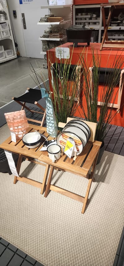This image depicts a display of outdoor furniture and tableware in a retail setting, likely in Gwangmyeong, South Korea. The scene is set on a light-colored, textured carpet.
In the foreground, a wooden folding table is set up. On the table, there are several items:
* A stack of plates featuring a hot dog design, with a yellow "SALE" tag attached.
* A white ceramic mug with a black rim.
* A white bowl with a black rim.
* A clear glass vase with a light blue, patterned design.
* A light pink, patterned bag with white circles.
To the left of the table, a black fabric and wooden folding chair is visible. Behind the table and chair, two tall, artificial plants with thin, grass-like leaves are placed.
In the background, a red display unit can be seen, with a black cast-iron grill on top. Shelving units containing various items, possibly kitchenware or decorative objects, are also visible on the shelves behind the red unit. A closed door with a handle is present on the far left of the image, and some additional shelving units with boxed items are to its right. The environment suggests a home goods or furniture store.