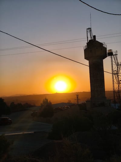 This image captures a sunset in Riba-roja de Túria, Spain, with a prominent water tower silhouetted against the bright sun. The sky transitions from a deep blue at the top to a warm orange near the horizon. In the foreground, a road curves through a sparsely vegetated area, with a red car parked on the left. Several trees dot the landscape, some appearing silhouetted. A series of power lines stretch across the sky. To the right, the tall, concrete water tower stands with an antenna on top, accompanied by a complex metal structure. Further in the background, rolling hills and distant structures are visible, softened by the sunset's glow. The overall scene conveys a sense of twilight stillness in an industrial or semi-rural setting.