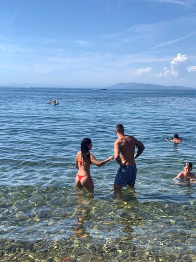 This media file depicts a sun-drenched day at Pozzarello, Italy, with several individuals enjoying the shallow, clear waters of a lake or sea. The sky is a brilliant blue, with wispy white clouds, suggesting a pleasant, warm day. The water is transparent, revealing a bed of pebbles beneath its surface, which is rippled by a gentle breeze.
In the foreground, a man and a woman are interacting. The man, with a muscular build and visible tattoos, wears dark patterned swim trunks. The woman is in a red bikini. They appear to be in a playful or affectionate stance. Further out in the water, other people are visible. Two young boys are wading or swimming, their faces turned towards the viewer. Another person is visible further out, appearing to swim or float. In the distance, a small group is on a paddleboard.
The scene is one of relaxation and recreation. The bright sunlight casts reflections on the water, highlighting the textures of both the surface and the submerged pebbles. The overall impression is of a lively summer day at the beach or lake, with people enjoying the cool water and the sunshine. No visible text is present in the image.