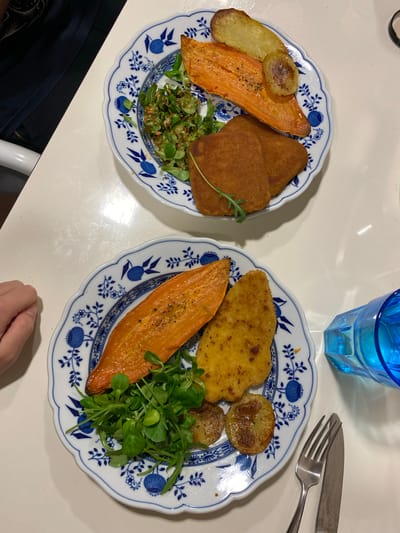 Two plates of food are arranged on a white surface. Each plate has a blue and white floral pattern around the rim. The plate on the left contains two rectangular pieces of golden-brown food, a slice of cooked sweet potato, a potato slice, and some greenery. The plate on the right contains a piece of golden-brown food, a slice of cooked sweet potato, several potato slices, and greenery. A blue glass, a fork, and a knife are located near the right plate. A person's hand is partially visible near the bottom right of the image. The environment appears to be indoors, likely a dining area. The location is not identifiable as Villaviciosa, Spain from this image.