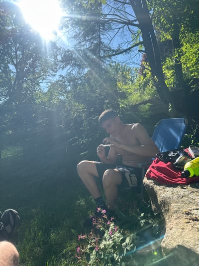 The image depicts an outdoor scene in Panagyurski kolonii, Bulgaria, under bright daylight conditions. A young adult male with light skin and short dark hair is seated shirtless on a blue collapsible camping chair, wearing dark shorts with a white Nike logo and dark athletic shoes with patterned socks. He is actively engaged in handling a small white object, possibly rolling paper or a cigarette, with both hands, looking downwards.
To the right of the seated male, a large, light-colored rock or boulder serves as a makeshift table. Upon this rock, several items are placed: a folded red piece of clothing, a black smartphone or similar electronic device, multiple small rectangular packages (possibly cigarette boxes or similar items), and a yellow plastic bottle with a black cap.
The environment is characterized by dense green foliage, including numerous trees with visible trunks and branches, and lush green grass. Small pink-purple flowering plants are visible in the foreground near the bottom right. The sky, visible through gaps in the tree canopy, is clear blue. Direct sunlight from the upper left quadrant of the frame causes significant lens flare and bright illumination across the scene, indicating clear weather. In the bottom left corner, a partial view of a foot wearing a dark athletic shoe and dark socks is visible, suggesting the presence of another individual, possibly the photographer.