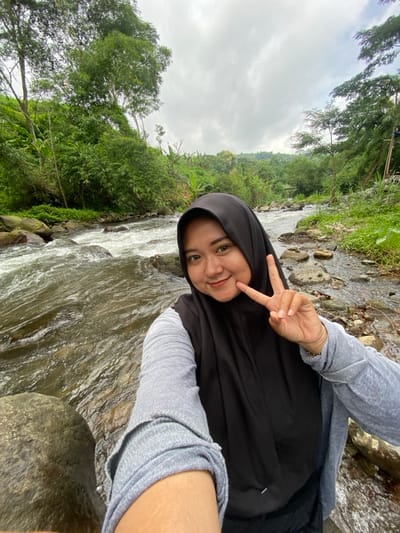 A woman wearing a black hijab and grey shirt makes a peace sign while sitting beside a rushing river. Green trees and foliage line the riverbank under a cloudy sky. The photo is taken at an angle, with the river flowing diagonally across the frame. A large rock is visible in the foreground.