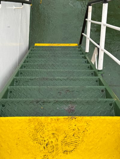 This media file depicts a close-up view of a metal staircase with diamond plate treads, painted a dark green. The staircase is situated on what appears to be a deck, with a section of bright yellow painted surface to its right. A distinct shoe print is visible on the yellow surface, suggesting recent activity. To the left of the staircase, a wet, green-colored deck surface is visible, with a white railing system above it. The deck appears to be outdoors, as indicated by the wetness and the overall industrial setting. The lighting suggests it could be daytime, but the presence of water on the deck indicates it might be raining or has recently rained. The location is identified as Mers El Hadjadj, Algeria. There is no visible text in the image. The overall impression is one of an industrial or maritime setting, possibly a ferry or a port facility, given the sturdy construction of the staircase and the presence of safety railings. The focus on the textures of the painted metal and the shoe print suggests an emphasis on the functional and worn aspects of the environment.