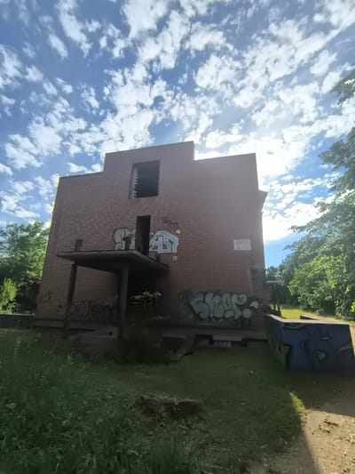 The image shows an abandoned brick building with graffiti covering its exterior. The building has several large, dark windows, some of which appear broken or boarded up. A small, covered porch extends from the front of the building. The surrounding area is overgrown with grass and surrounded by trees. The sky is blue with scattered white clouds, and the sun is shining brightly, suggesting it is daytime. The overall scene evokes a sense of neglect and decay. There is a sign visible on the right side of the building that appears to be a warning or informational plaque, but the text is too small to read clearly. There is also graffiti on the lower parts of the building and on a concrete structure in the foreground. The location is identified as Rannamõisa, Estonia.