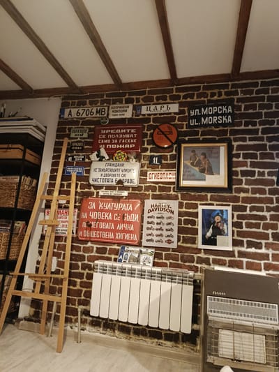 A cluttered interior space in Bryastovets, Bulgaria, features a brick wall adorned with various signs and memorabilia, including license plates, street signs in Bulgarian and English (e.g., "UL. MORSKA," "A 6706 MB"), safety signs, and Harley-Davidson advertisements. A wooden easel leans against the wall, near a white radiator and a gas heater. Framed images are visible, including one of a musician with a violin. A shelf with wicker baskets holding various items stands to the left.