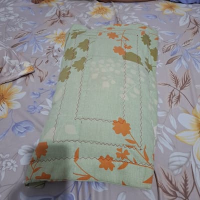 The image displays a light green pillow with an orange floral pattern, lying on a floral patterned bedsheet in Reudeuep, Indonesia. The pillow has visible stitching in red thread, creating a square border pattern. The bedsheet features a repeating motif of large yellow and white flowers with brown and blue leaves. The lighting suggests an indoor setting, possibly a bedroom, with the bed occupying the majority of the frame. No people or specific actions are depicted.