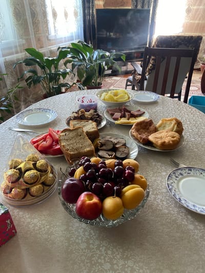 A table is set with a meal, including fruit, bread, meat, cheese, and fried dough. The table is covered with a patterned tablecloth. In the background, there is a television, a chair, and plants. The scene appears to be indoors, likely a home dining area, with natural light coming from a window.