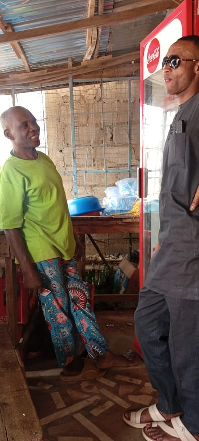 Two men are standing in an indoor space in Kwachiri, Nigeria. The older man is wearing a light green t-shirt, patterned pants, and sandals. He is leaning against a crate. The second man is wearing a dark gray shirt and pants, sunglasses, and white sandals. He is standing next to a Coca-Cola refrigerator. Behind them is a caged wall and a wooden table with bottled water and other items. The floor has a geometric pattern. The roof structure is visible, made of wood and metal sheeting.