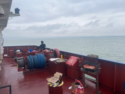 The image displays the red-painted deck of a vessel, captured on a body of water consistent with the coastal areas near Bangor, United Kingdom, such as the Menai Strait. The sky is uniformly overcast with grey clouds, and distant landmasses, including a faint industrial-type structure, are visible on the horizon. One individual, clad in a dark hooded garment, stands with their back to the observer, positioned near a large marine winch spooled with blue rope. To the starboard side of the deck, a black freestanding barbecue grill actively cooks multiple sausages and other meat cuts. A silver metal tray rests on the blue railing directly above the grill, and a partially open brown paper bag, labeled "FULZ" or "FUELS," sits on the deck nearby. A red emergency bunker station box is affixed to a bulkhead. Further forward, a small cylindrical stand supports an open cardboard box and an empty dark glass bottle, with yellow and black hazard tape wrapped around the stand. The water surface is calm.