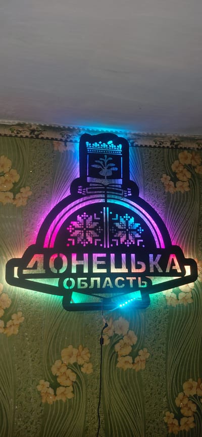 An illuminated metal sign, likely for the Donetsk Oblast, is mounted on a wall with floral wallpaper. The sign features a decorative top with what appears to be a crown and a stylized plant or tree. Below this, a circular element contains intricate geometric patterns resembling snowflakes. The text "ДОНЕЦЬКА" (Donetsk) is prominently displayed in large letters, with "ОБЛАСТЬ" (Oblast) written below it in a smaller font. The sign is illuminated by colorful LED lights, cycling through shades of blue, pink, and green. A black wire extends from the sign down the wall. The wallpaper is green with a textured pattern and scattered yellow floral motifs. The image is taken from a slightly low angle, looking up at the sign. There are no people visible in the image. The location is described as Ketrysanivka, Ukraine.