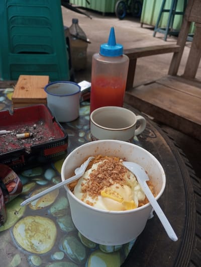 The image captures an outdoor dining scene in Gadog, Indonesia. In the foreground, a white paper bowl filled with instant noodles, topped with a fried egg and fried shallots, sits on a table covered by a patterned tablecloth depicting light-colored stones. Two white plastic forks are inserted into the noodles. To the left of the bowl, a red plastic ashtray contains multiple cigarette butts and ash. Adjacent to the ashtray are a small wooden block and a white enamel mug with a blue rim. Behind these items, a translucent plastic squeeze bottle with a blue cap, containing a red sauce, stands upright. To the right of the sauce bottle and behind the noodle bowl is a cream-colored ceramic mug with a handle. The background shows an unpaved ground surface. On the left, a green plastic chair is visible, while on the right, a wooden chair with a slatted backrest is present. Further in the background, a large plastic water bottle and green corrugated metal structures are discernible, with a bicycle wheel featuring a blue rim partially visible near the structures. No individuals are present in the scene.
