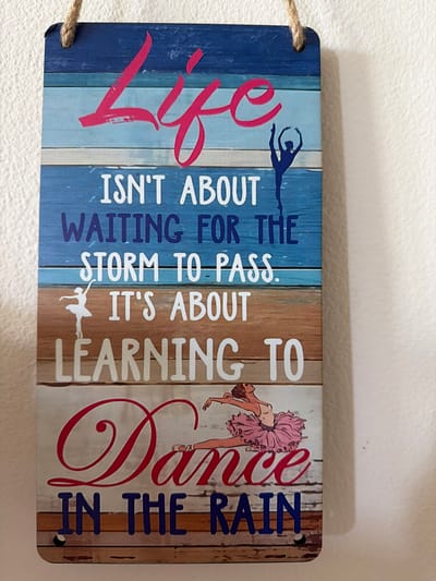 A rectangular wooden sign with a distressed wood plank design hangs from a piece of jute rope against a white, textured wall. The sign features a motivational quote about life and dancing. The quote reads: "Life isn't about waiting for the storm to pass. It's about learning to dance in the rain." The word "Life" is written in large, stylized pink script at the top. Below "Life," in white sans-serif font, are the words "ISN'T ABOUT WAITING FOR THE STORM TO PASS." and "IT'S ABOUT LEARNING TO". The word "Dance" is prominently displayed in large, swirling red script in the lower half of the sign, with the words "IN THE RAIN" written below it in bold blue sans-serif font. The sign also includes silhouette illustrations of ballerinas: one in a dynamic pose at the top, and another in a graceful pose in a pink tutu in the lower half. The image is a close-up, slightly angled shot of the sign, with the wall providing the background. There is no indication of a city or country in the image itself.