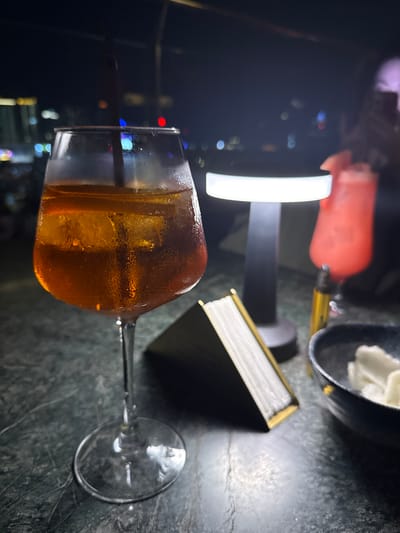 A close-up, slightly blurry shot shows a glass filled with an amber-colored liquid and ice, resting on a dark, speckled surface, likely a table. A straw is visible within the glass. In the background, out of focus, is a modern, brightly lit lamp with a white cylindrical shade, and a glimpse of a pinkish-red drink in another glass or container. To the right of the main drink, a small, open notebook with gold-edged pages is visible. The overall ambiance suggests a nighttime setting, possibly in a bar or restaurant, with blurred lights indicating a city view in the distance. The dimly lit scene and the presence of drinks imply a social gathering or a moment of relaxation. The location context is Khan Boeng Keng Kang, Cambodia.