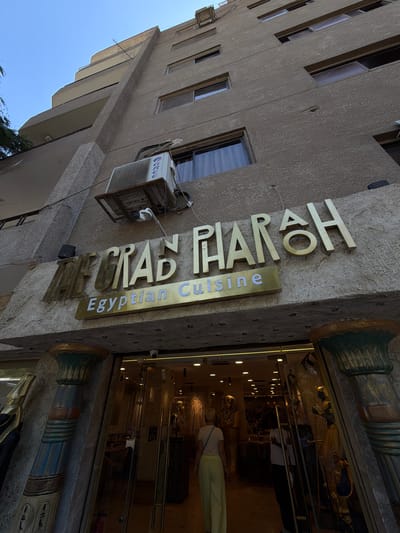 This is a tilted, eye-level, exterior shot of a building in Kafrat Nassar, Egypt. The building has a rough, textured, beige facade. There are multiple windows visible on the building, some with dark frames and others with lighter frames. An air conditioning unit is mounted on the side of the building.

A prominent sign made of gold-colored, raised lettering is attached to the building. The sign reads "THE GRAND PHARAOH" vertically, with "Egyptian Cuisine" written horizontally on a gold plaque below. The letters are three-dimensional and appear to be metallic.

To the right of the sign, an entrance to a building is visible. It is brightly lit from within and appears to be a store or restaurant, with display cases showcasing Egyptian-themed artifacts, including sarcophagi and statues. A person wearing a white top and light-colored pants is visible inside, looking at the displays. The entrance is flanked by decorative columns that have an Egyptian architectural style, featuring hieroglyphics and motifs.

The lighting suggests it is daytime, with natural sunlight illuminating the scene. The sky is a clear blue, indicating fair weather. The overall impression is one of a commercial establishment with an ancient Egyptian theme.