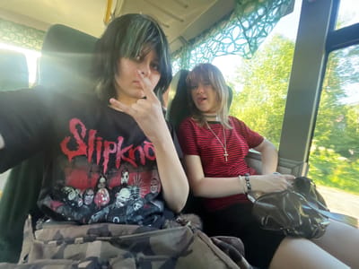 Two young individuals are seated next to each other inside a vehicle, possibly a bus or train, with a blurred background of trees and foliage visible through the window. The individual on the left, with dark hair and green highlights, is wearing a black t-shirt with a graphic print that appears to be band merchandise. This person is making a hand gesture with their right hand, forming a rock-and-roll sign. They also have a nose ring. The individual on the right has lighter colored hair and is wearing a red and black striped shirt. They are looking towards the camera with their tongue sticking out slightly. They are wearing a choker necklace with a pendant that resembles an ankh cross. A dark-colored handbag is placed on their lap. The seating and window structure suggest they are in transit. The location of Kurtukovo, Russia, is not discernible from the visual elements within the image itself.